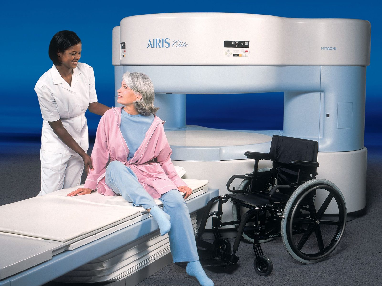 A technologist standing beside a patient seated on the table of a Hitachi AIRIS Elite open MRI, with a wheelchair alongside.