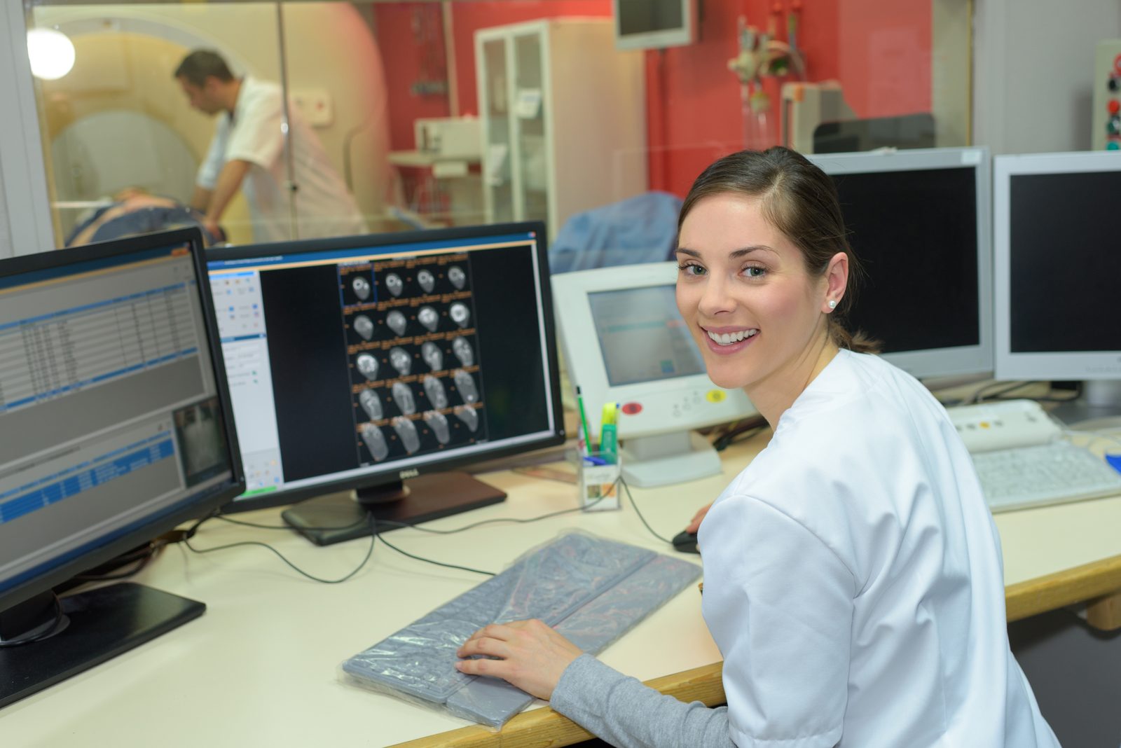 A technologist at the imaging console reviewing scan images, with the scan room visible through the window.